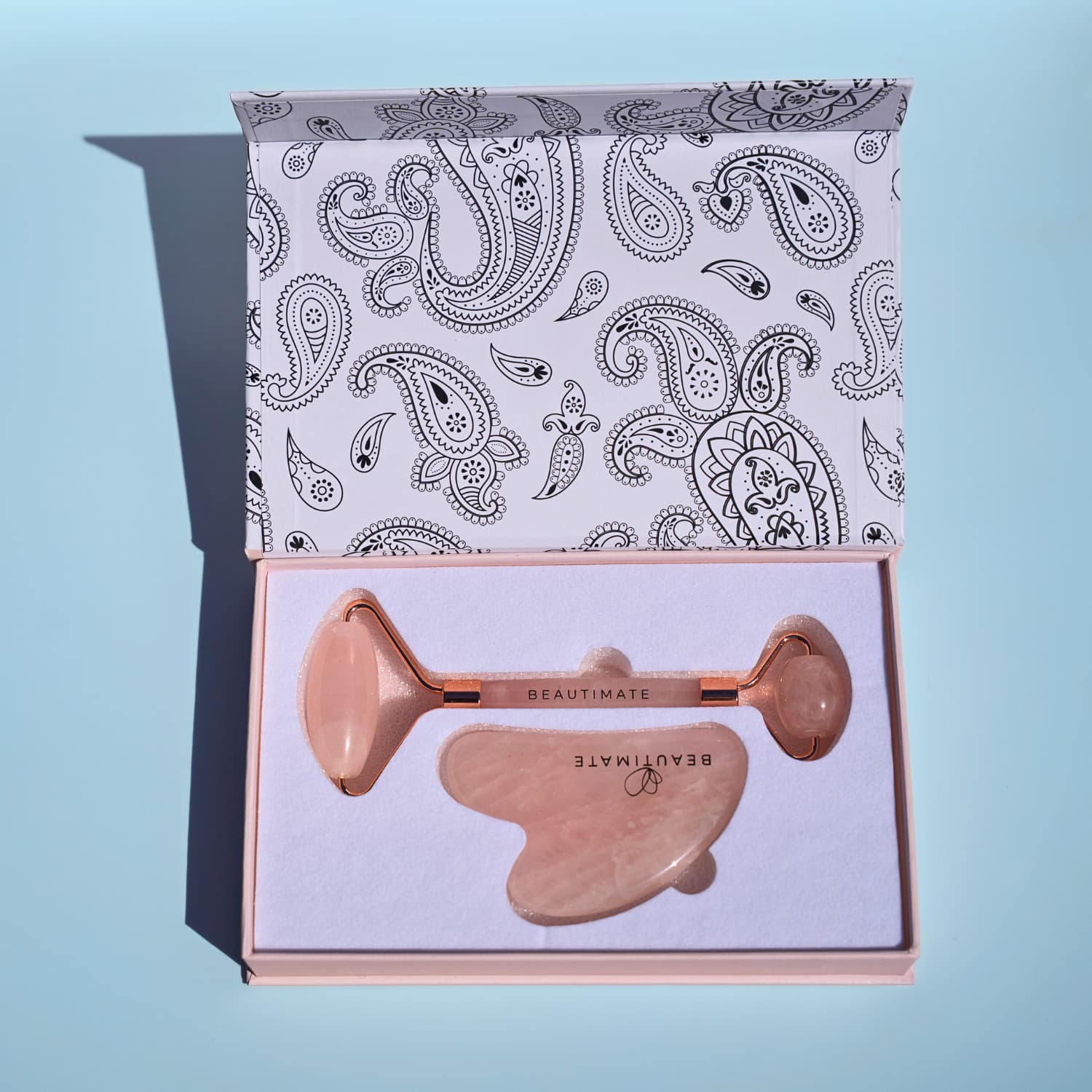 Rose gold facial roller with paisley patterned box on a light blue background